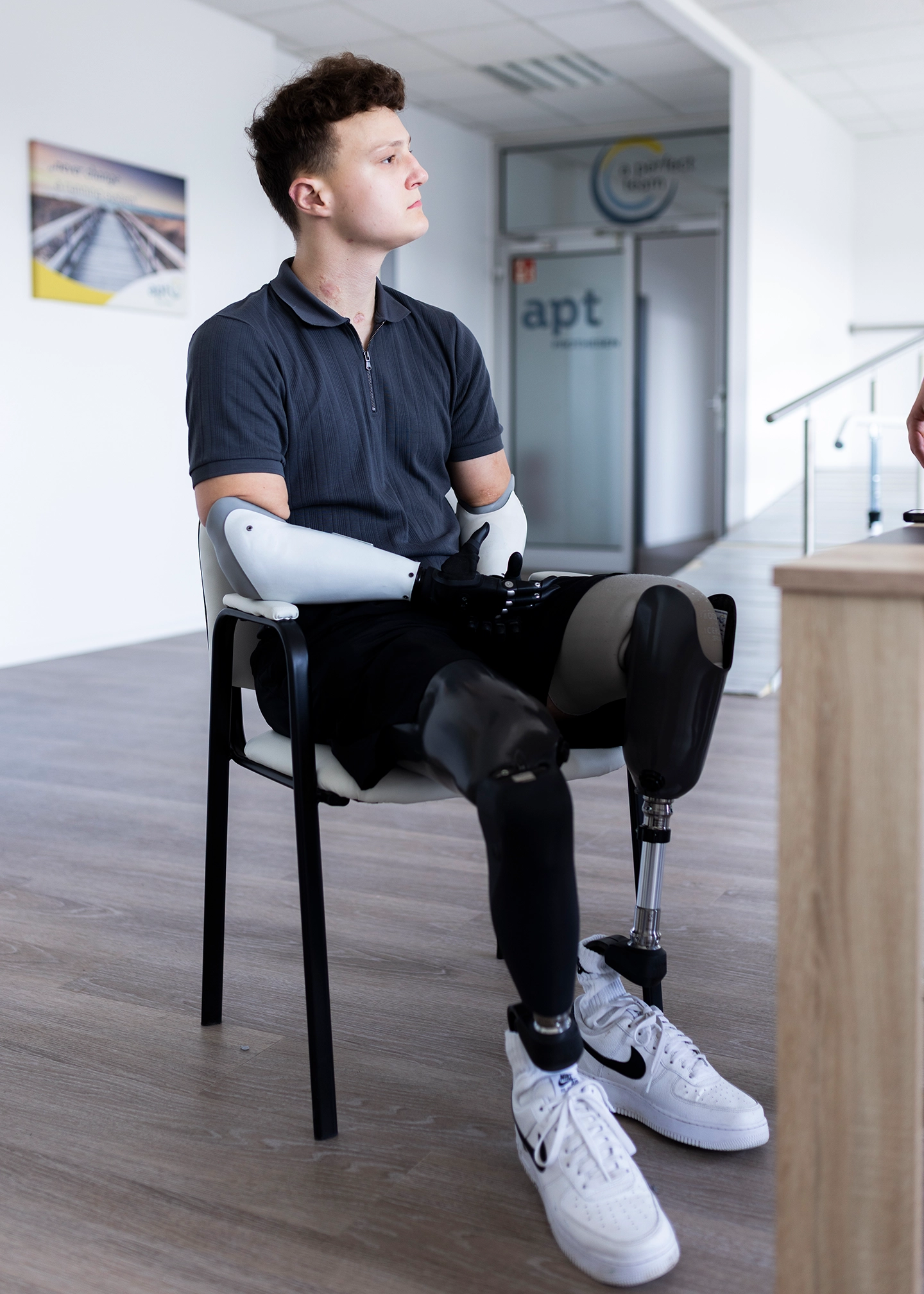 Junger Mann mtizwei Armprothesen und zwei Beinprothesen sitzt auf einem stuhl in einer APT Prothesen Filiale und schaut nach rechts
