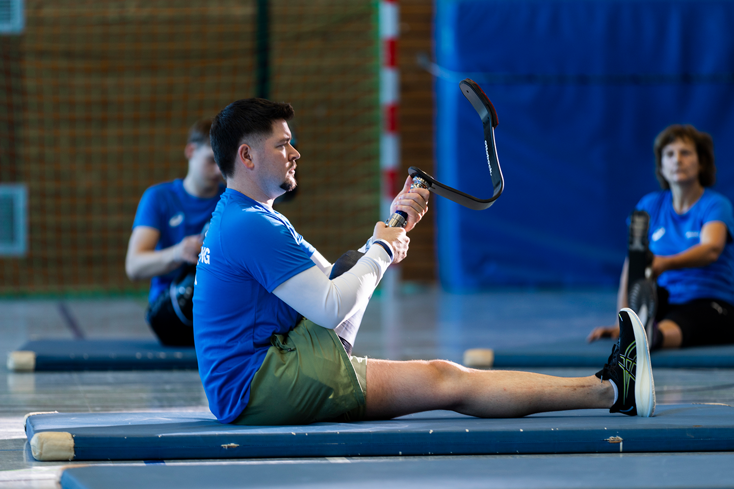 Träger einer Beinprothese sitzt in der sporthalle auf einer blauen matte und hält seite Sportprothese nach oben
