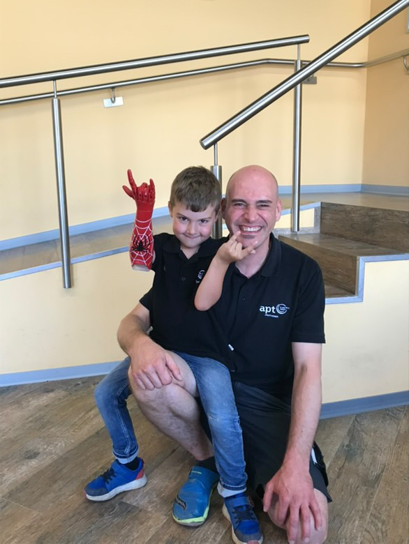 APT-Mitarbeiter Alexander Paul mit einem Prothesenträger Kind auf dem Schos, Kind trägt eine Armprothese im Spiderman Look