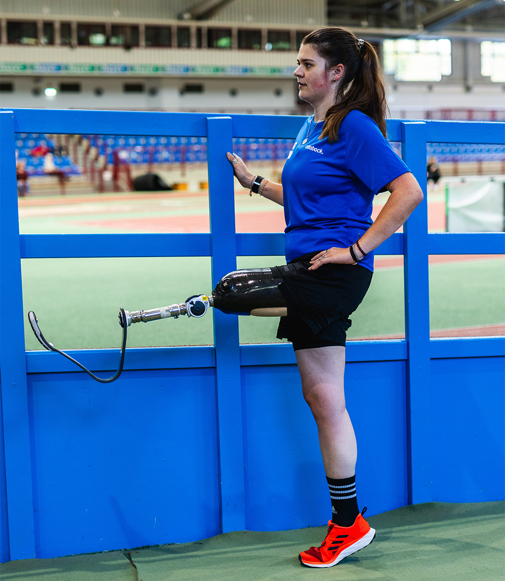 APT Prothesen Kundin Lisa Seidel stützt ihre Sportprothese auf einem Zaun am Sportplatz