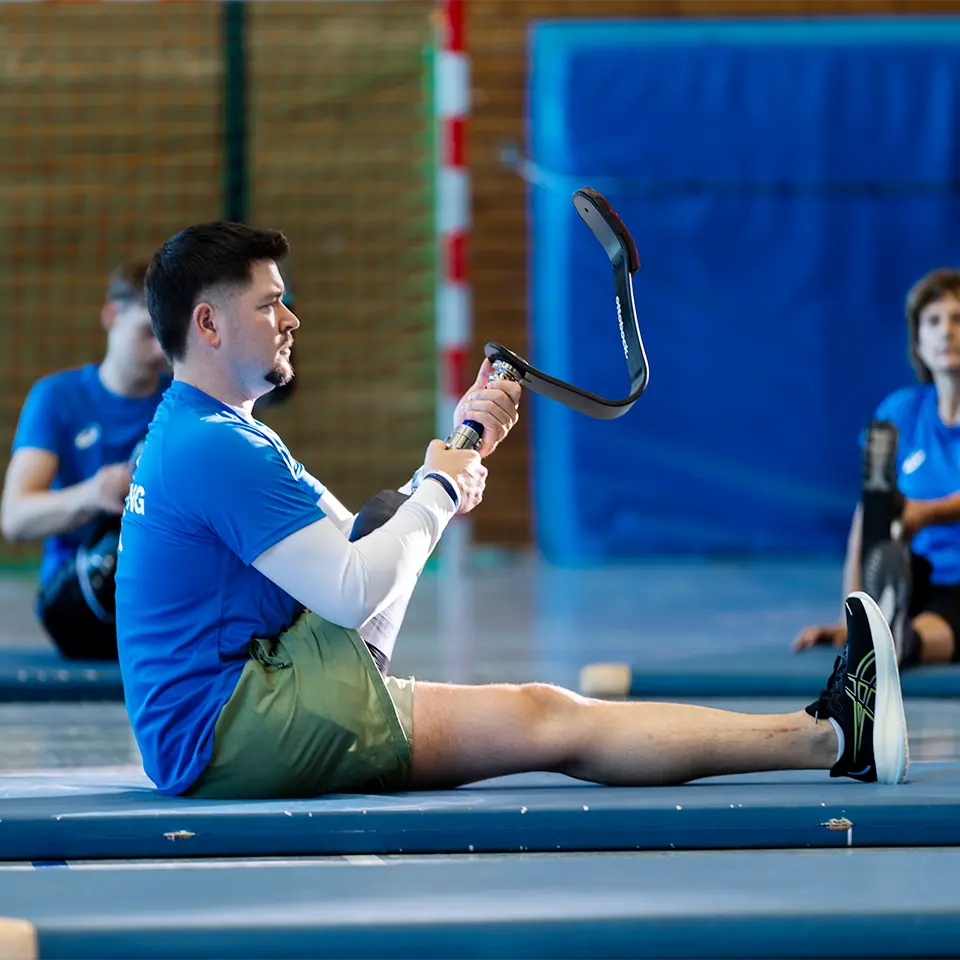 Mann in Sportklamottten sitzt in einer Turmhalle und dehnt sich mit seiner Sportprothese von APT Prothesen.