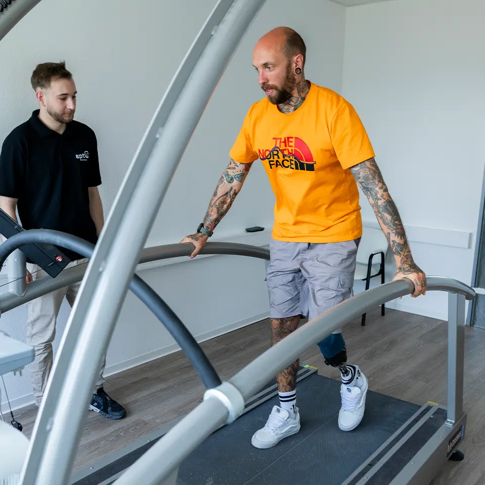 Ein Mann mit Beinprothese steht auf einem Laufband und stützt sich auf den Trägern des Laufbands ab. Er trainiert mit der Begleitung eines APT Mitarbeiters das Laufen mit seiner neuen Beinprothese.